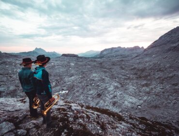 Zwei Personen mit Hüten und Blasinstrumenten stehen auf einem felsigen Gelände und blicken auf eine weite Berglandschaft.