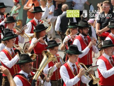 Eine Blaskapelle in traditioneller österreichischer Tracht marschiert in einer Parade mit Schaulustigen im Hintergrund.