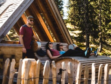 Drei Personen entspannen sich auf Holzliegen vor einer rustikalen Hütte, umgeben von Bäumen und Sonnenlicht.