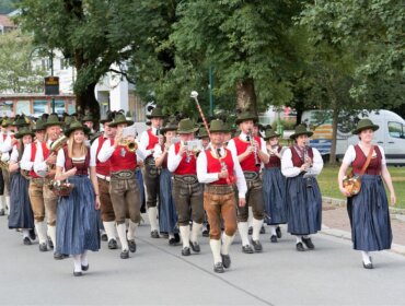 Ein Spielmannszug in traditioneller österreichischer Tracht spielt im Freien auf einer Straße, die von Bäumen und Gebäuden gesäumt ist.