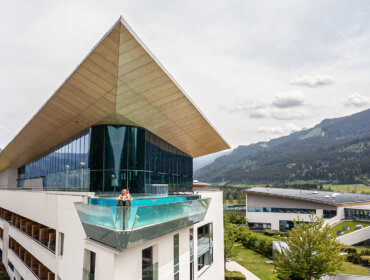 Eine Person steht auf einem Glasbalkon eines modernen Gebäudes mit österreichische Bergen und Grün im Hintergrund.