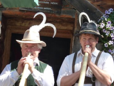 Zwei Männer in traditioneller österreichischer Tracht spielen Alphörner vor einem rustikalen, mit Blumen geschmückten Holzgebäude.