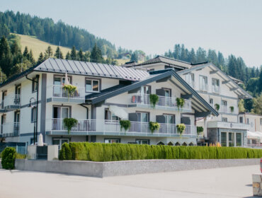 Ein modernes alpines Hotel mit Balkonen und Grünflächen, eingebettet in einen bewaldeten Berghang unter einem klaren Himmel.