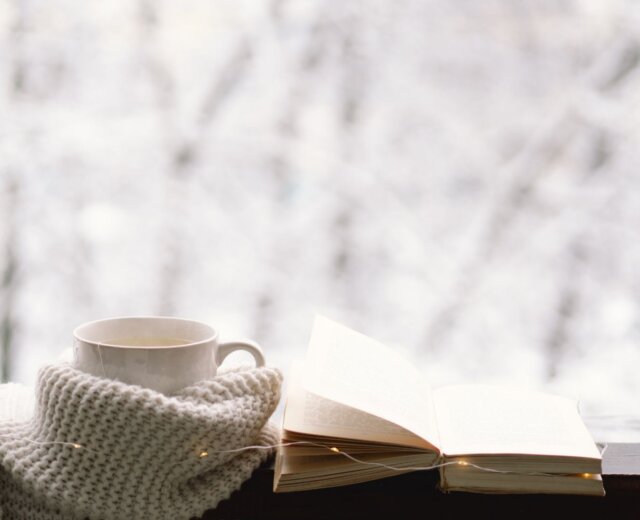 Eine Tasse und ein aufgeschlagenes Buch, eingewickelt in einen gestrickten Schal, auf einer Fensterbank vor einem verschneiten Hintergrund.