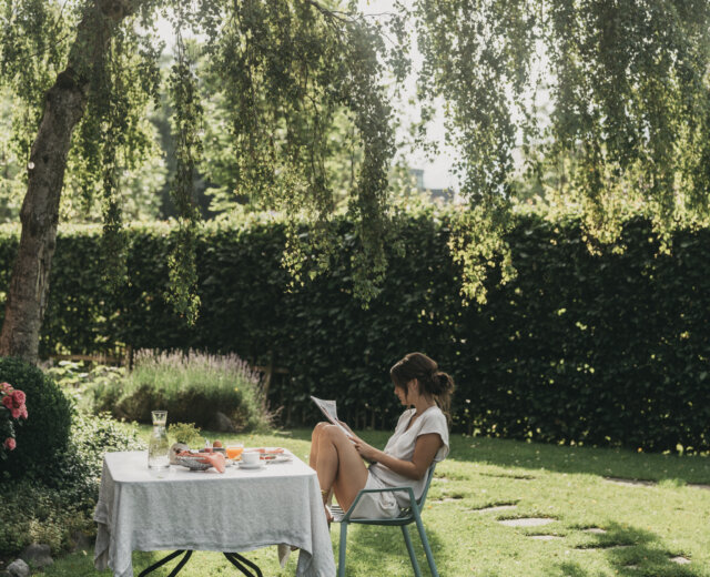 Eine Frau liest an einem gedeckten Tisch mit Speisen und Getränken in einem sonnenbeschienenen Garten unter einem belaubten Baum.