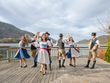 Sechs Personen in traditioneller österreichischer Kleidung tanzen im Freien auf einem Holzdeck an einem See und in den Bergen.