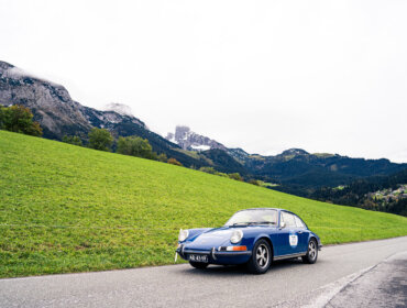 Ein klassisches blaues Auto fährt auf einer Straße mit grünen Hügeln und Bergen im Hintergrund.