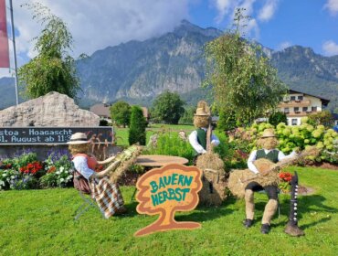 Drei Strohfiguren in traditioneller Kleidung sitzen neben einem Bauernherbst-Schild in einem malerischen österreichischen Bergdorfgarten.