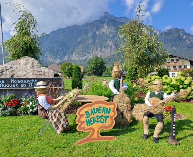 Drei Strohfiguren in traditioneller Kleidung sitzen neben einem Bauernherbst-Schild in einem malerischen österreichischen Bergdorfgarten.