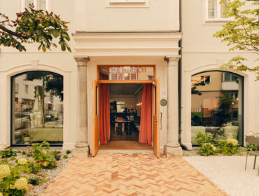 Cremefarbener Gebäudeeingang mit offenen Holztüren, gewölbten Fenstern und Sitzgelegenheiten im Freien mit Grünpflanzen.