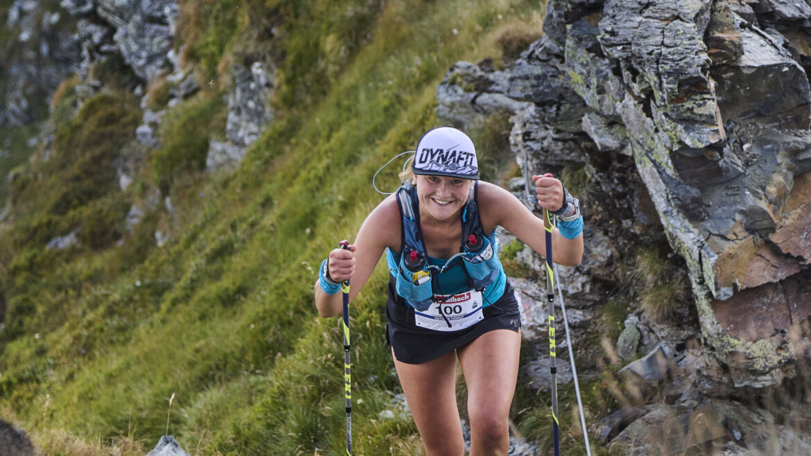 Eine Frau wandert mit Trekkingstöcken einen felsigen Bergpfad hinauf, umgeben von steilen, grasbewachsenen Hängen. (vergrößerte Ansicht)