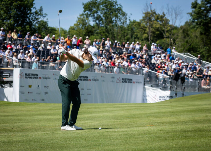 Ein Golfer schwingt den Ball auf einem grünen Platz, während eine große Menschenmenge von der Tribüne aus bei sonnigem Himmel zusieht.