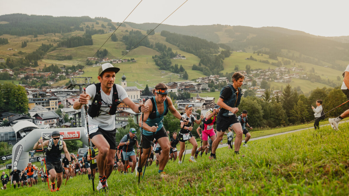Eine Gruppe von Läufern wetteifert bergauf auf einem grasbewachsenen Hang mit einer Stadt und Bergen im Hintergrund. (vergrößerte Ansicht)