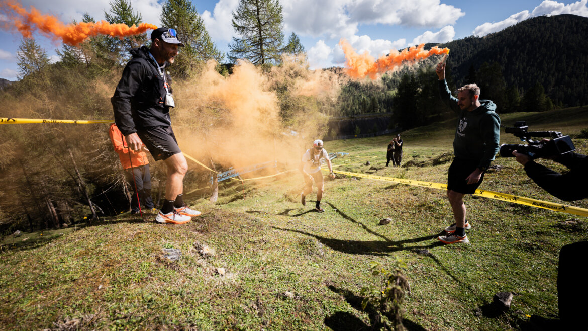 Zwei Männer mit orangefarbenen Rauchfackeln feuern einen Läufer während eines Rennens in einer malerischen Berglandschaft an. (vergrößerte Ansicht)
