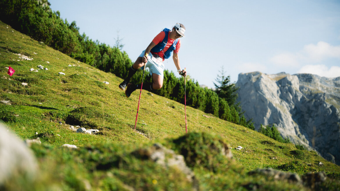 Eine Person, die in den Bergen wandert, springt mit Hilfe von Trekkingstöcken auf einem grasbewachsenen Abhang in die Luft. (vergrößerte Ansicht)