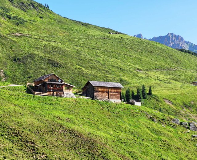 Zwei Holzhütten stehen an einem üppig grünen Hang mit Bergen im Hintergrund unter einem klaren blauen Himmel.