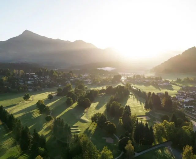 Ein sonnenbeschienenes Tal mit Bäumen, Häusern und Bergen im Hintergrund bei Sonnenaufgang.