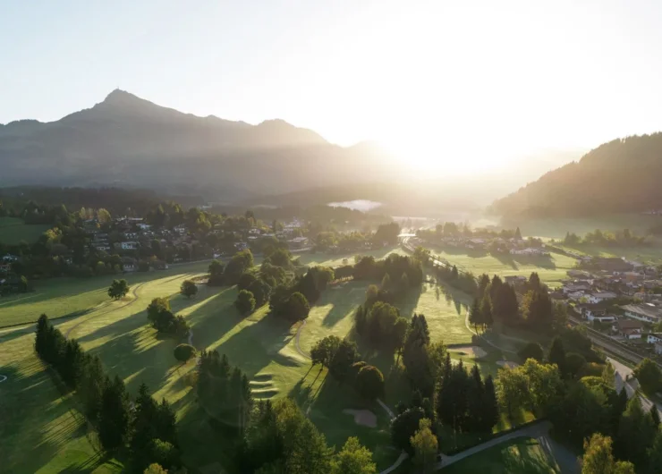 Ein sonnenbeschienenes Tal mit Bäumen, Häusern und Bergen im Hintergrund bei Sonnenaufgang.