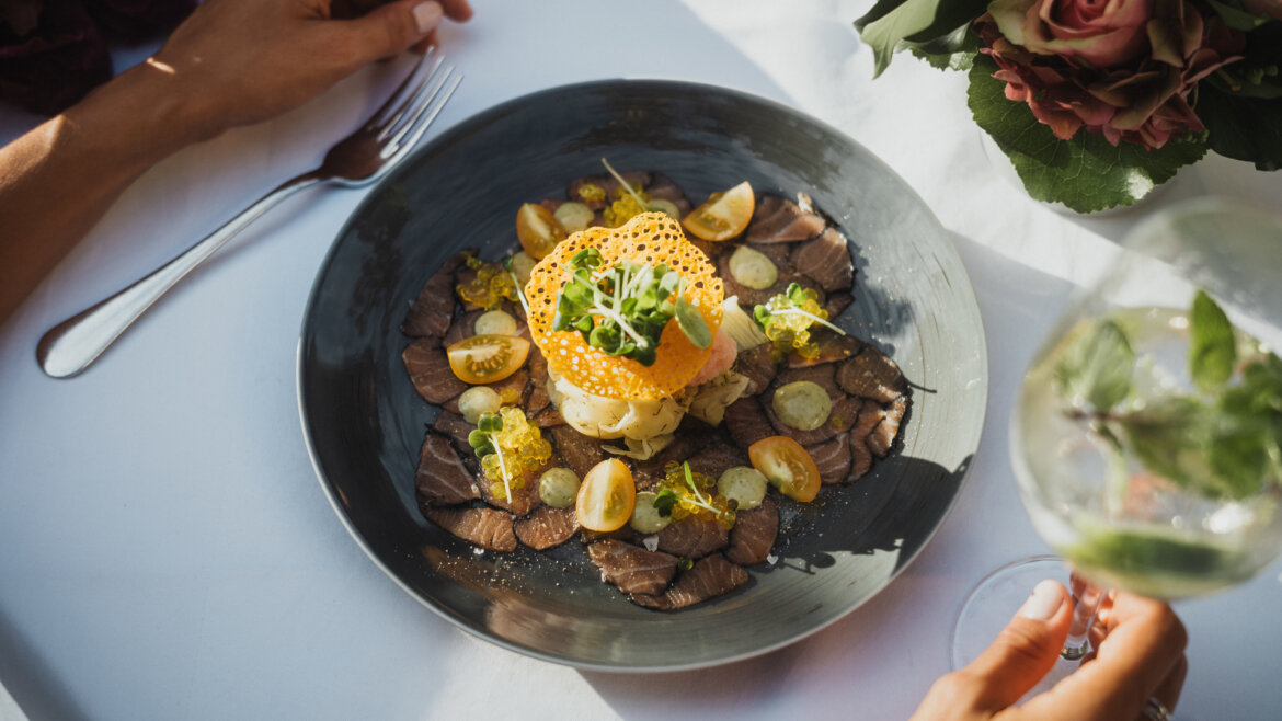 Ein Gourmet-Gericht mit geschnittenem Fleisch, Soße, Tomaten und Beilagen auf einem schwarzen Teller, mit Händen und einem Glas daneben. (vergrößerte Ansicht)
