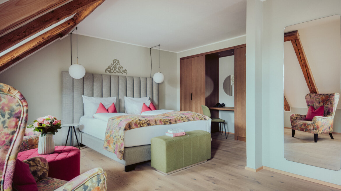 Helles Schlafzimmer im Dachgeschoss mit Blumendekor, großem Bett, Sessel, Spiegel und Holzakzenten. (vergrößerte Ansicht)