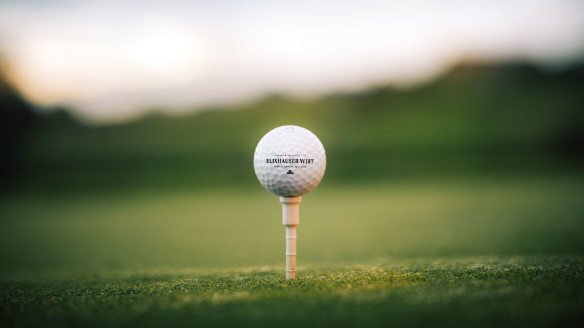 Ein Golfball auf einem Abschlag sitzt auf einem grasbewachsenen Platz, mit einem unscharfen grünen Hintergrund. (vergrößerte Ansicht)