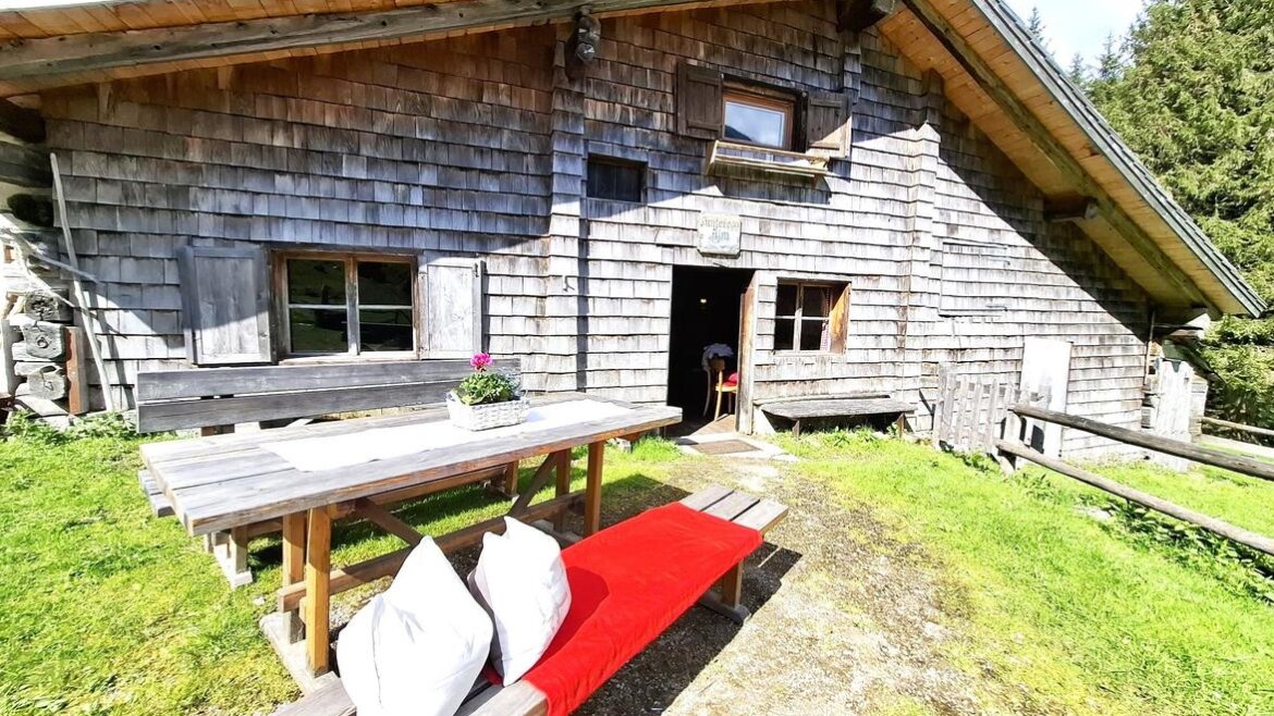 Österreichische Holzhütte mit Picknicktisch, roter Bank und weißen Kissen draußen, umgeben von Bäumen und Sonnenlicht.