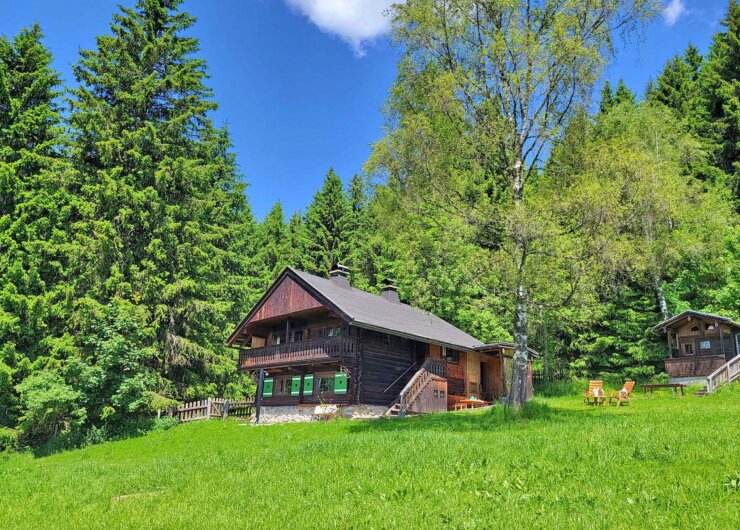 Eine Holzhütte, umgeben von grünem Gras und hohen Bäumen unter einem strahlend blauen Himmel mit vereinzelten Wolken.