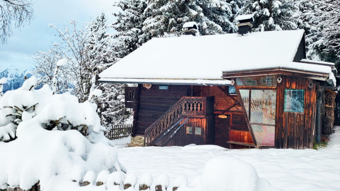 Eine schneebedeckte Holzhütte steht zwischen verschneiten Bäumen und einem Holzzaun in einer Winterlandschaft. (vergrößerte Ansicht)