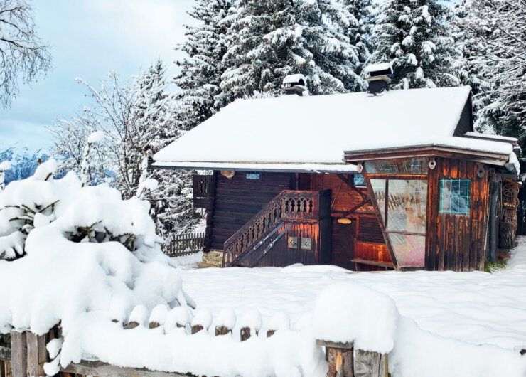 Eine schneebedeckte Holzhütte steht zwischen verschneiten Bäumen und einem Holzzaun in einer Winterlandschaft.
