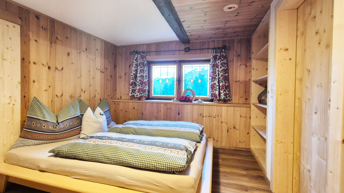 Gemütliches Holzschlafzimmer mit Doppelbett, kariertem Bettzeug, Fenster mit geblümten Vorhängen und eingebauten Regalen. (vergrößerte Ansicht)
