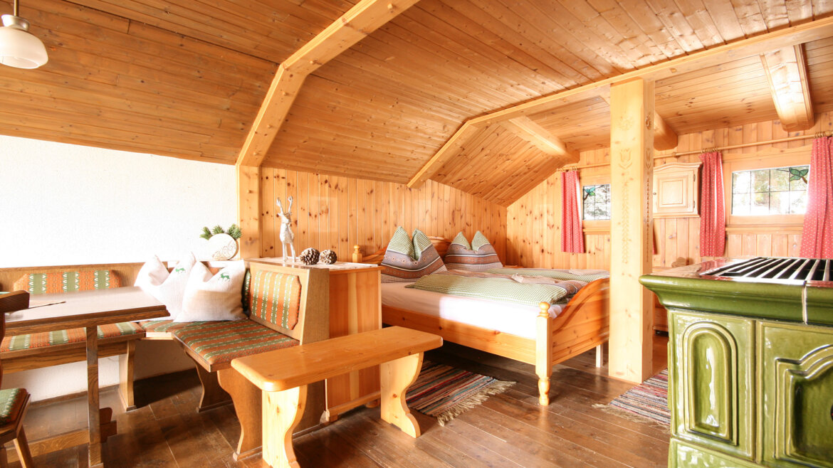 Gemütliche Holzhütte mit Bett, Essecke, Bänken und rustikaler Einrichtung in warmem Tageslicht. (vergrößerte Ansicht)