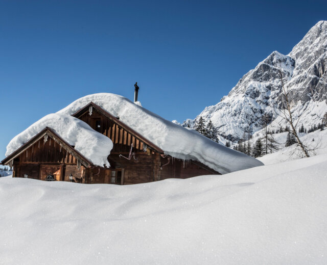 Winterlandschaft Eine schneebedeckte Holzhütte liegt vor verschneiten Bergen und einem strahlend blauen Himmel.
