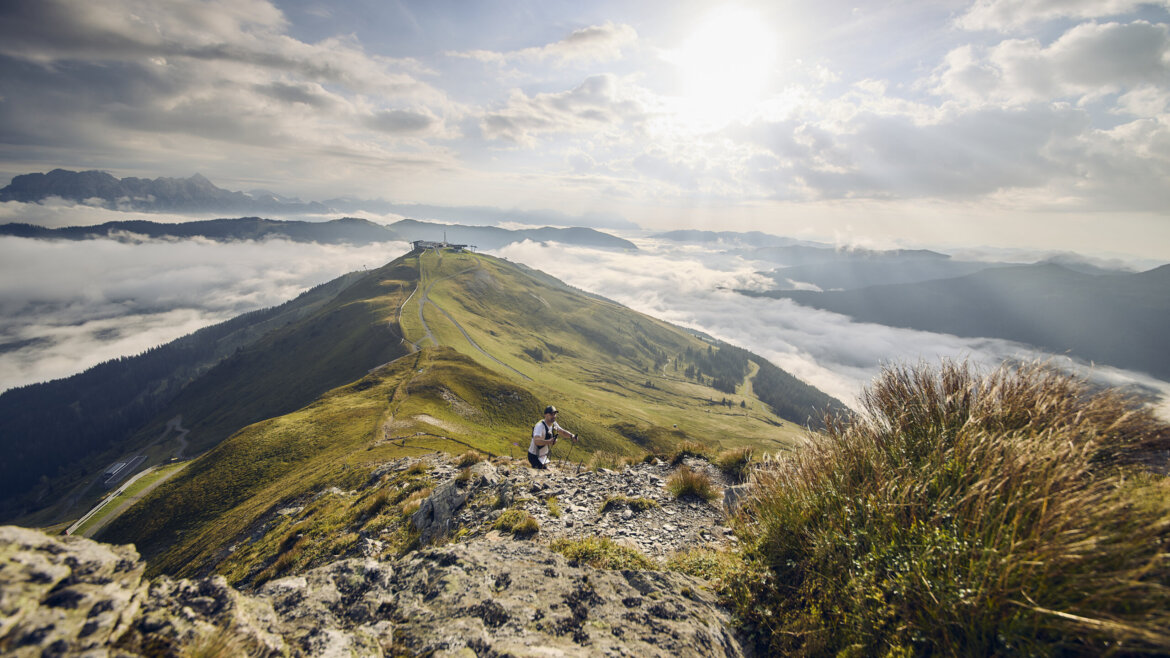 Ein Wanderer erklimmt bei strahlendem Sonnenschein einen felsigen Gebirgskamm, mit Wolken und sanften Hügeln im Hintergrund. (vergrößerte Ansicht)