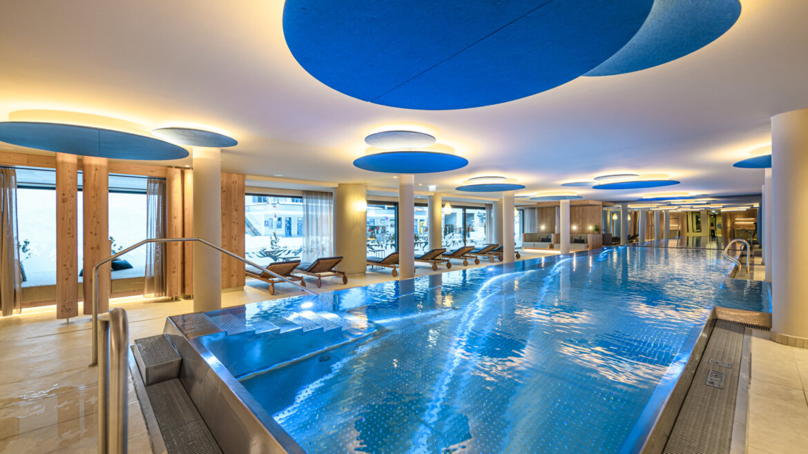 Modernes Hallenbad mit blauen runden Deckenleuchten und Liegestühlen an großen Fenstern. (vergrößerte Ansicht)