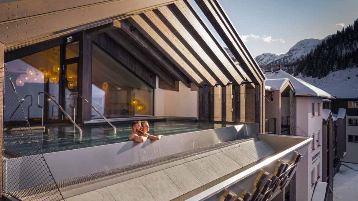 Ein Paar genießt den Außenpool auf dem Dach eines modernen Hotels mit Blick auf die Berge. (vergrößerte Ansicht)