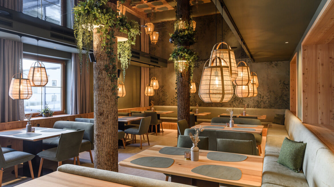 Modernes Restaurant mit Holztischen, grünen Stühlen, großen Hängelampen und Baumstammdekor. (vergrößerte Ansicht)