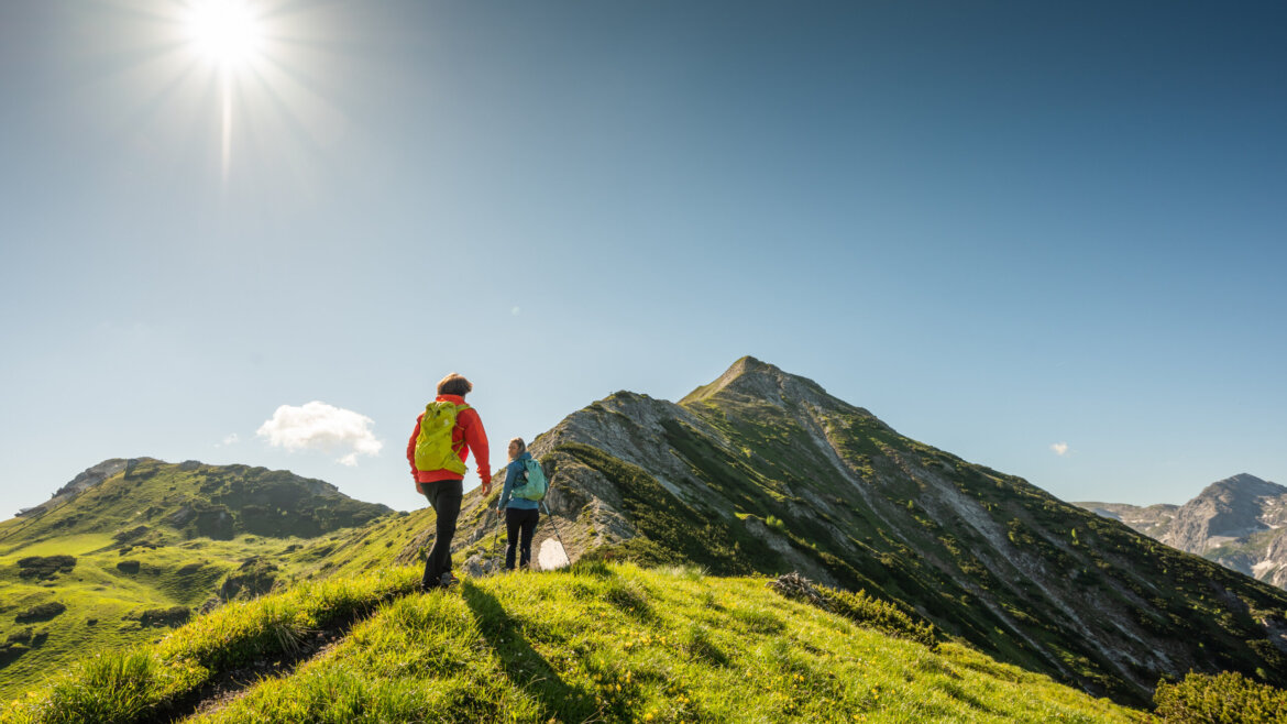 Zwei Wanderer stehen auf einem grasbewachsenen Bergrücken unter strahlender Sonne und blauem Himmel. (vergrößerte Ansicht)