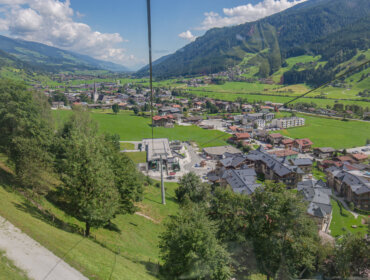 Ein malerisches Bergdorf mit grünen Feldern, Häusern und fernen Bergen unter einem teilweise bewölkten Himmel.