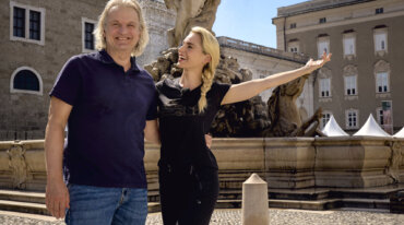 Ein Mann und eine Frau lächeln an einem sonnigen Tag an einem historischen Brunnen, zu sehen in Almsommer im TV.