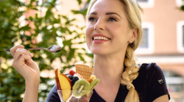 Lächelnde Frau mit geflochtenem Haar genießt einen Fruchteisbecher in einem Café im Freien - Almsommer im TV-Flair.