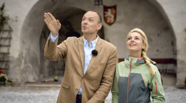 Ein Mann und eine Frau stehen draußen, lächelnd unter einem steinernen Torbogen - Almsommer im TV hat es perfekt eingefangen.