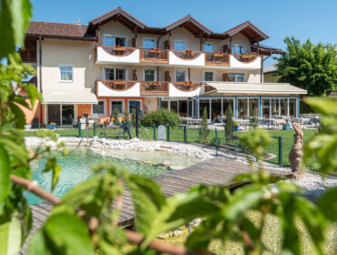Sonniges Hotel mit Balkonen mit Blick auf einen kleinen Teich und eine Holzbrücke, umrahmt von grünen Ästen.
