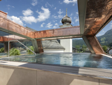 Pool auf dem Dach mit Metallrahmen, Blick auf die Berge und den Kirchturm unter einem blauen Himmel mit Wolken.