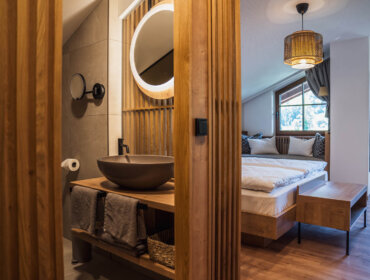 Modernes Schlafzimmer mit Holzdekor, einem runden Spiegel über einem Waschbecken und einem ordentlich gemachten Bett am Fenster.