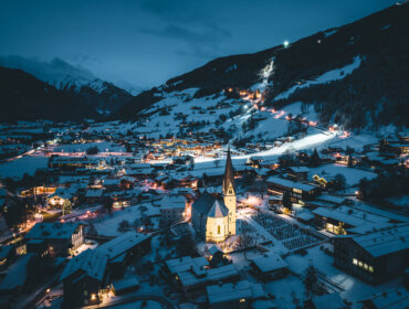 Ein verschneites Dorf in der Abenddämmerung, die Kirche in der Mitte, mit beleuchteter Skipiste und Berghäusern im Hintergrund.