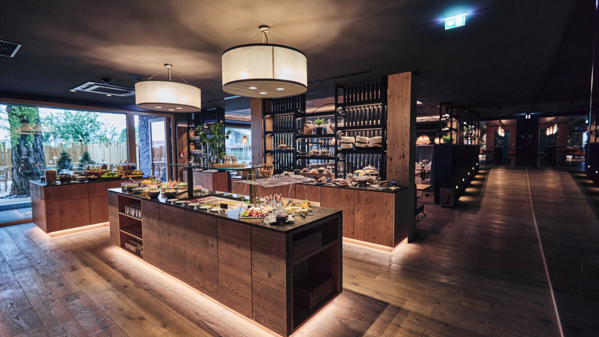 Modernes Buffetrestaurant mit Holzdekor, beleuchteten Theken und verschiedenen Speisenauslagen auf zentralen Tischen. (vergrößerte Ansicht)