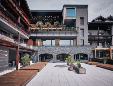 Moderner Hotelinnenhof mit Steinmauern, Holzbalkonen und Topfpflanzen auf einer großen gepflasterten Terrasse.