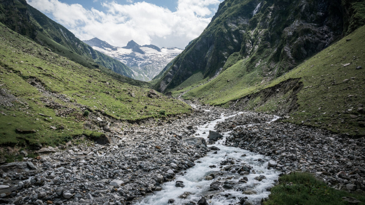 Ein felsiger Gebirgsbach fließt durch ein grünes Tal mit schneebedeckten Gipfeln im Hintergrund unter einem blauen Himmel. (vergrößerte Ansicht)
