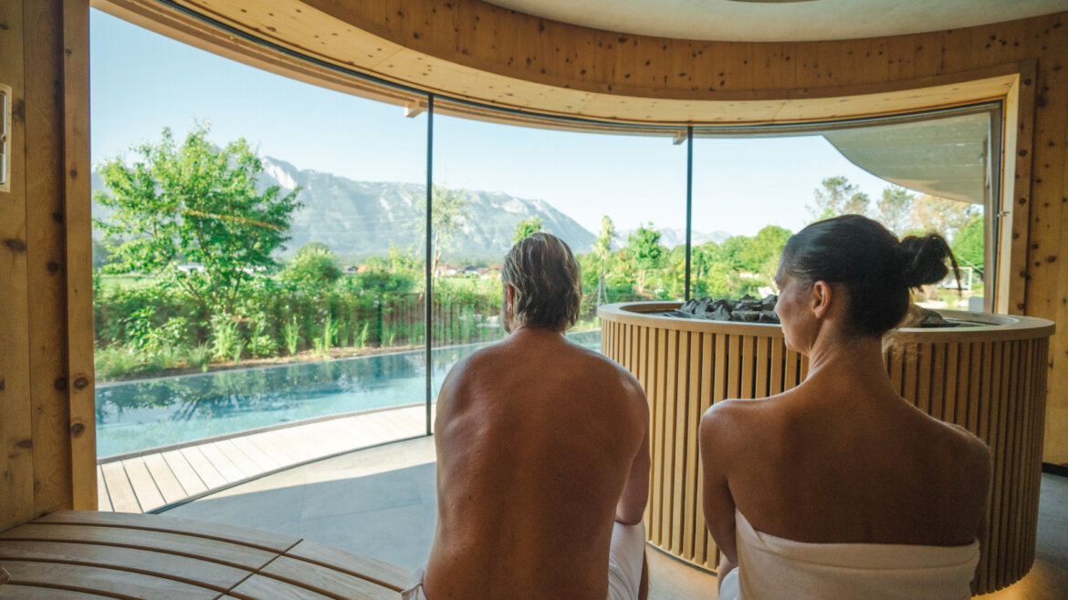 Zwei Personen in Handtüchern sitzen in einer Sauna mit großen Fenstern und Blick auf die Berge und den Pool. (vergrößerte Ansicht)
