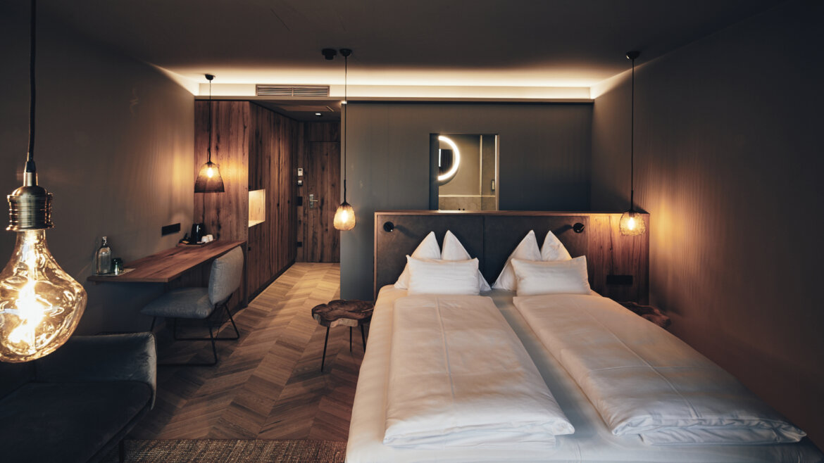 Modernes Hotelzimmer mit Doppelbett, Hängelampen, Holzschreibtisch und offenem Bad in warmer Beleuchtung. (vergrößerte Ansicht)